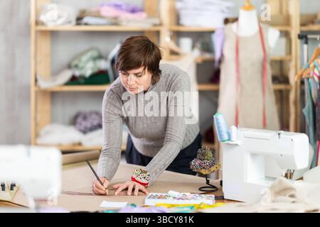 Die Schneiderin zeichnet in der Nähwerkstatt anhand von Mustern eine Skizze der zukünftigen Kleidung auf Papier Stockfoto