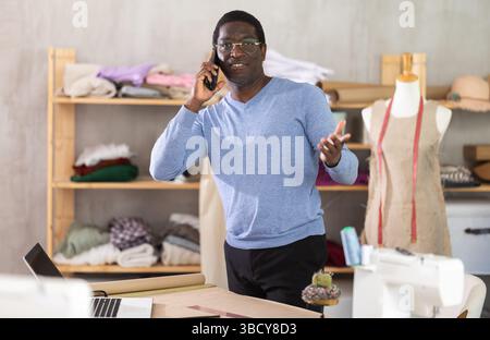 Männliche Schneiderin, die telefoniert und den Laptop im Nähstudio überprüft Stockfoto