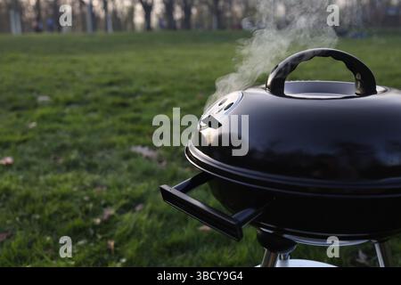 Nahaufnahme eines schwarzen runden Holzkohlegrills mit Dampf aus dem Deckel auf grünem Gras in einem Park Stockfoto