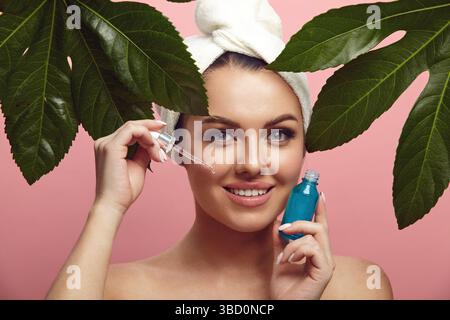 Lächelnde schöne junge Frau im Kopftuch mit Gesichtsserum steht zwischen tropischen Blättern über rosa isoliertem Hintergrund. Werbung für Schönheit Stockfoto