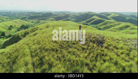 Aus der Vogelperspektive eines Touristen, der auf einem Hügel mit offenen Armen steht und die atemberaubende Landschaft der Insel Sumba, Indonesien, mit üppigem grünem Gras und sanften Hügeln bedeckt ist Stockfoto