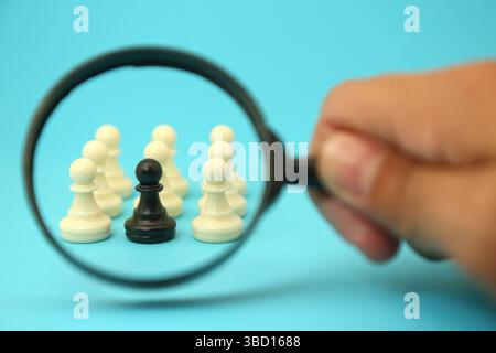 Ein einzelner schwarzer Bauer unter Beobachtung, hervorgehoben durch eine Lupe, inmitten einer Gruppe weißer Bauer. Konzept bei der Auswahl von Mitarbeitern Stockfoto
