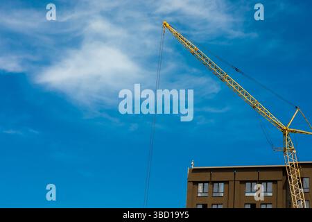 Gelber Baukran, der sich über einem modernen Gebäude unter einem leuchtend blauen Himmel mit schroffen Wolken erstreckt. Stockfoto