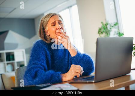 Ältere Frau im blauen Pullover gähnt beim Arbeiten am Laptop in einer gemütlichen, modernen Home Office-Umgebung Stockfoto