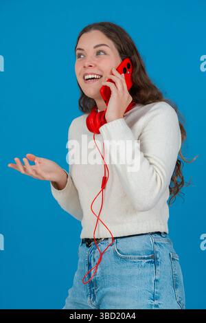 Vertikales Porträt einer freudigen Frau, die auf rotem Smartphone mit Kopfhörern spricht Stockfoto