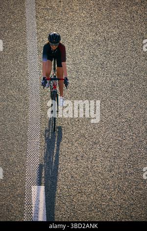 Blick von oben auf eine Radfahrerin, die allein auf einer leeren, strukturierten Autobahn fährt und einen langen Schatten entlang der gestrichelten Fahrbahnmarkierung wirft. Stockfoto