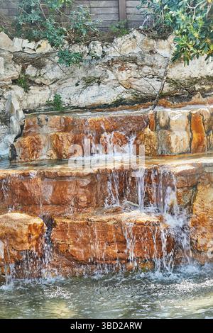 Das Wasser stürzt durch rostige rote Felsen in einem kleinen natürlichen Wasserfall, umgeben von Stein und üppig grünem Laub Stockfoto