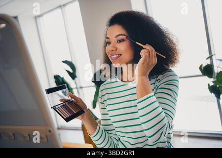Junge Frau, die in einem gemütlichen Wohnzimmer mit Tageslicht und natürlichem Licht Make-up aufträgt, um den Komfort des Wohnhauses zu demonstrieren Stockfoto