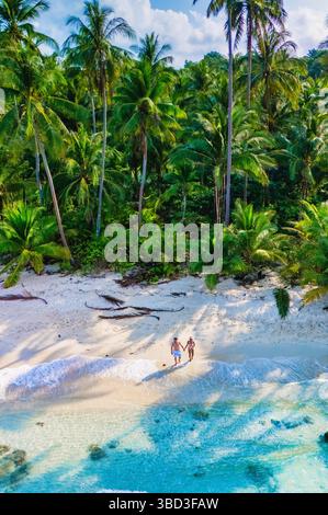 Goldener Sand und klares türkisfarbenes Wasser umschließen ein Paar, das Hand in Hand an einem ruhigen Strand geht. Hoch aufragende Palmen bilden eine malerische Kulisse von Koh Kood Thailand Stockfoto
