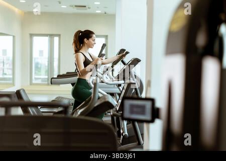 Eine Frau genießt ihre Fitness-Routine auf einem Crosstrainer, konzentriert und motiviert, umgeben von eleganten Fitnessgeräten bei hellem Tageslicht. Stockfoto