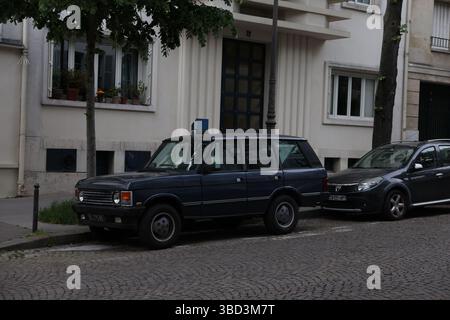 Zwei Autos – ein dunkelblauer Range Rover und ein dunkelgrauer Renault – parken auf einer Kopfsteinpflasterstraße vor einem weißen Gebäude mit einer tiefblauen Tür. Stockfoto