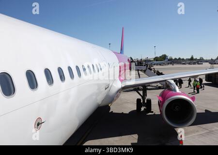 Auf dem Rollfeld des Flughafens parkt ein kommerzielles Flugzeug mit markantem rosafarbenem Schwanz und Motor, während die Passagiere über eine mobile Treppe einsteigen. Das geschäftige Ai Stockfoto