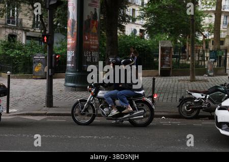 Zwei Fahrer auf einem Motorrad navigieren durch eine geschäftige Stadtstraße, beide tragen Helme zur Sicherheit. Die Szene zeigt ein geparktes Motorrad, ein rotes Pedal Stockfoto