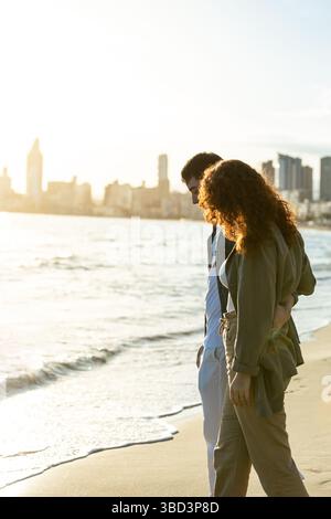 Junges Paar, das Arm in Arm am Strand bei Sonnenuntergang geht, einen zarten Moment mit sanftem Sonnenlicht und der Skyline der Stadt in der Ferne, die sich auf dem reflektiert Stockfoto