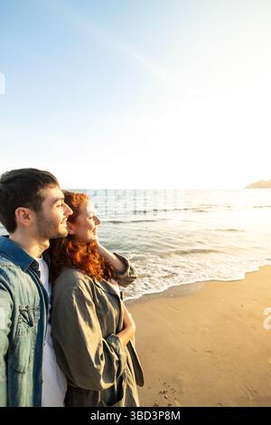 Glückliches Paar, das in der Nähe an einem Sandstrand steht und in den glühenden Sonnenuntergang über dem Meer blickt und einen ruhigen und liebevollen Moment im goldenen Li teilt Stockfoto