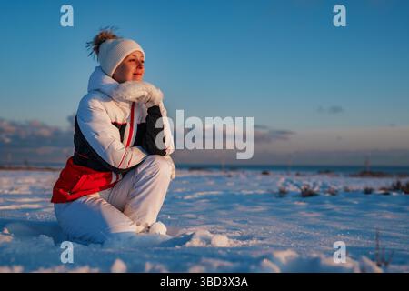 Glückliche junge Frau im Winteranzug sitzt im Schnee und blickt auf den Sonnenuntergang Stockfoto