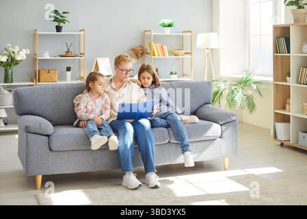 Großmutter und zwei Kinder sitzen auf der Couch, lesen zusammen Buch und genießen die Freizeit zu Hause. Stockfoto