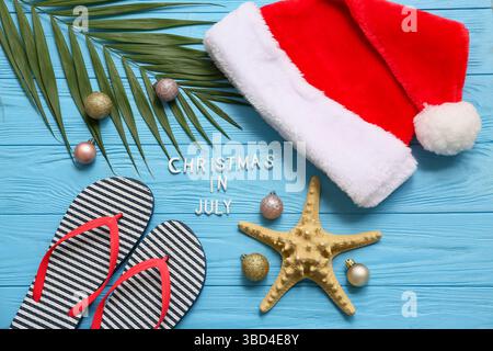 Weihnachtsmann Hut, Seesterne, Flipflops und Palmenzweig mit Weihnachtskugeln und Text WEIHNACHTEN IM JULI auf blauem Holzhintergrund. Draufsicht Stockfoto