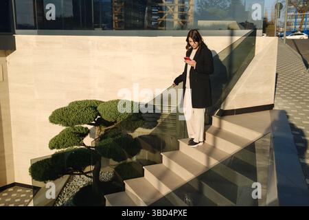 Professionelle Frau, die ein Smartphone benutzt, während sie Treppen in einem modernen Geschäftsgebäude hinabsteigt Stockfoto