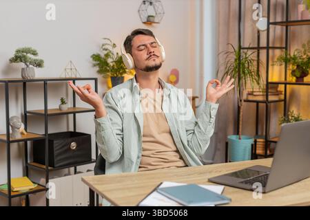 Beruhige dich, entspann dich. Der Mensch, der Musik hört, atmet tief, die Augen geschlossen, meditiert mit konzentrierten Gedanken, friedlichem Geist. Junger Freelancer sitzt am Arbeitsplatz des Home Office-Tisches. Lifestyle. Stockfoto