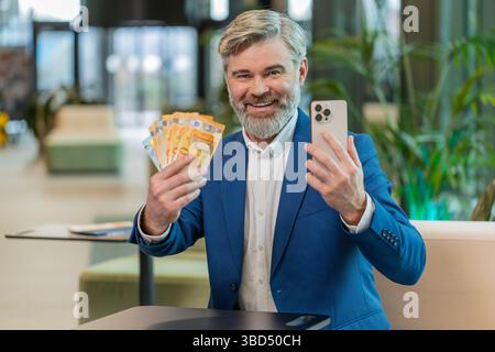 Erfolgreicher glücklicher reifer Geschäftsmann, der ein Smartphone hält und einen Fan von Euro-Rechnungen hält, der in der Lobby des Büros sitzt. Glücklicher kaukasischer bärtiger Kerl, der online Lotteriespiel gewinnt. Budget wird berechnet. Stockfoto