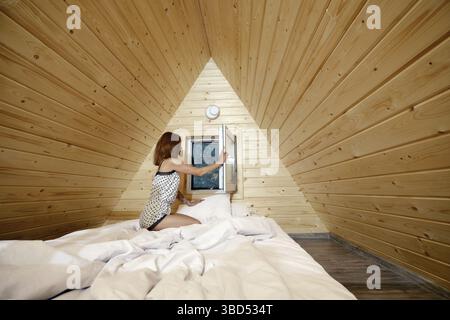 Frau im Pyjama öffnet das Fenster im Schlafzimmer der Blockhütte und genießt die frische Luft Stockfoto