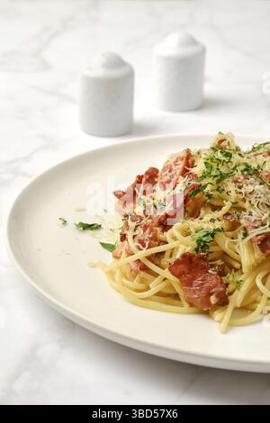 Aus nächster Nähe sehen Sie frisch zubereitete Spaghetti Carbonara mit knusprigen Speckstücken, geriebenem Käse und gehackten Kräutern auf einem glatten weißen Marmortisch Stockfoto