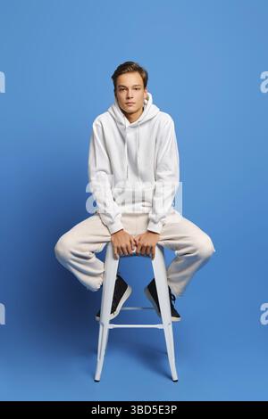 Hübscher Mann mit weißem Sweatshirt und Sweatshirts auf weißem Stuhl über blauem Studiohintergrund sitzend Stockfoto
