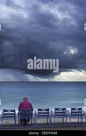 Ältere Mann auf Bank am Deich sitzen und beobachtete dunkle, bedrohliche Gewitterwolken über dem Meer Stockfoto