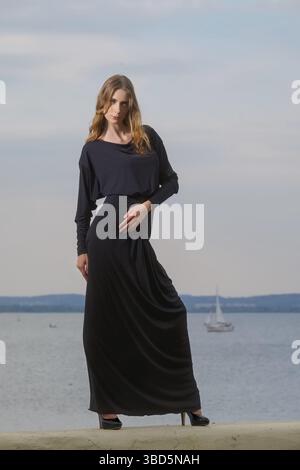 Attraktives Modemodell in langem schwarzem Kleid, das am Pier posiert Stockfoto