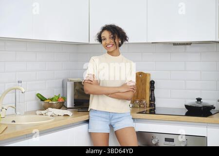 Junge lächelnde afroamerikanische Frau, die mit gekreuzten Händen in der Küche steht Stockfoto