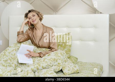 Nachdenkliche junge Frau sitzt im Bett mit einem Buch Stockfoto