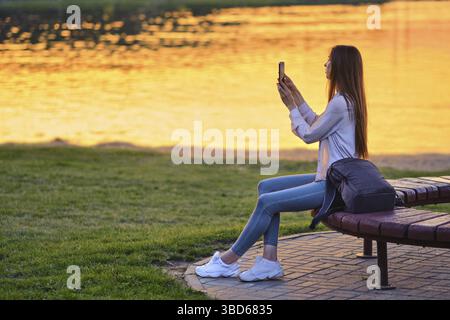 Bloggerin, die online den wunderschönen Sonnenuntergang streamt, sitzt auf einer Bank am Fluss Stockfoto