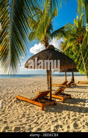 Sonnenliegen aus Holz, die von Sonnenschirmen mit Palmenblättern beschattet werden, an einem tropischen Sandstrand Stockfoto