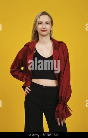 Eine junge Frau steht selbstbewusst vor einem leuchtend gelben Hintergrund und trägt ein markantes rotes Hemd über einem schwarzen Crop-Top. Ihre Pose vermittelt ein Gefühl für Stockfoto
