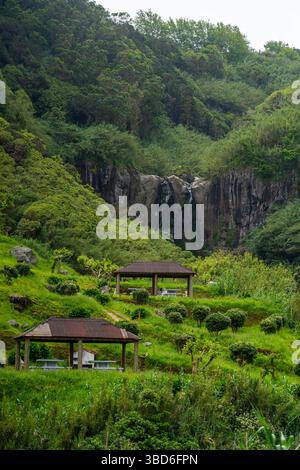 Rustikale Picknick-Unterstände umgeben von Grün unter einem kleinen Klippen-Wasserfall Stockfoto