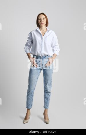 Entspannte Frau mittleren Alters mit weißem Hemd und blauer Jeans auf grauem Hintergrund Stockfoto