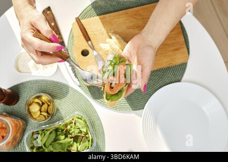 Von oben auf die Hände einer Frau mittleren Alters legt sie salzigen Lachs auf ein Sandwich, während sie Frühstück macht Stockfoto