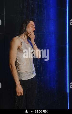 Ein langhaariger Mann im nassen T-Shirt steht in einer Dusche im Profil, beleuchtet durch Neonlicht Stockfoto