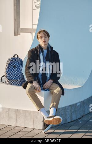 Junge Schülerin mit Rucksack in altem Wollmantel, Schal und kurzer Hose sitzt im Freien Stockfoto