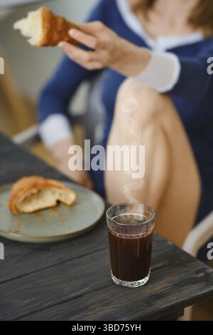 Unkenntliche Frau, die in der Küche mit Croissant in der Hand sitzt, konzentriert sich auf Glas mit Kaffee Stockfoto