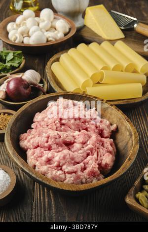Eine hölzerne Schüssel gefüllt mit rohem Hackfleisch ist umgeben von Pasta, Pilzen, Knoblauch und Käse auf einem rustikalen Holztisch, der eine lebhafte Auswahl an anderen bietet Stockfoto