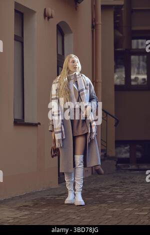Eine junge Frau in stilvollem Wollmantel, Schal und knielangen Stiefeln verließ das Haus und setzte ihr Gesicht den Sonnenstrahlen aus Stockfoto