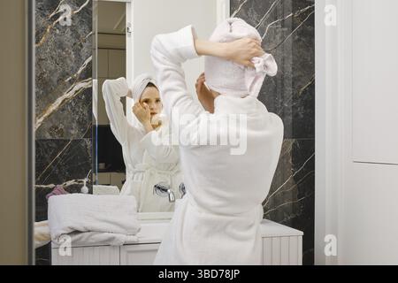 Frau im Bademantel, die nasse Haare mit einem Handtuch vor einem Spiegel umhüllt Stockfoto