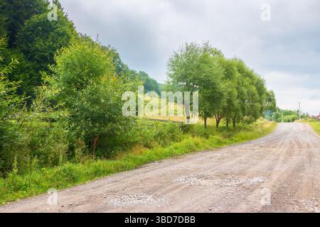 Schotterstraße führt durch die ländliche Landschaft von transkarpatien. Friedliche Landschaft auf dem Land zwischen bewaldeten Hügeln. Bewölkter Sommerhimmel. Bäume entlang der Stockfoto