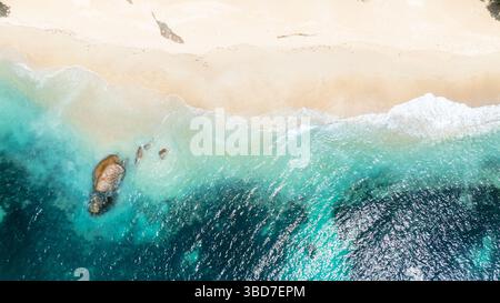 Aus der Vogelperspektive sehen Sie sanfte türkisfarbene Wellen, die die Sandküste mit einem Blick auf Felsen unter dem Wasser durchstreifen. Praslin, Seychellen. Stockfoto