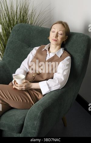 Frau mittleren Alters, die sich während der Kaffeepause mit geschlossenen Augen in einem Büroerholungsraum ausruht Stockfoto