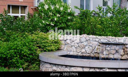 Dekorativer Teich mit Steinen in einem kleinen Garten. Blühender weißer Flieder. Kleiner Brunnen. Ein Innenhof in der Stadt. Stockfoto