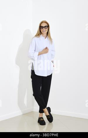 Mode-Porträt im urbanen Stil einer glücklichen lächelnden Frau mit weißem Hemd und schwarzer Hose. Fröhliche Frau, die mit überkreuzten Beinen in einem hellen Raum steht Stockfoto