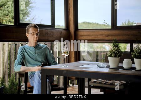 Ein Mann mittleren Alters sitzt auf der Veranda des Landhauses mit einem Glas Wein und benutzt ein Smartphone Stockfoto
